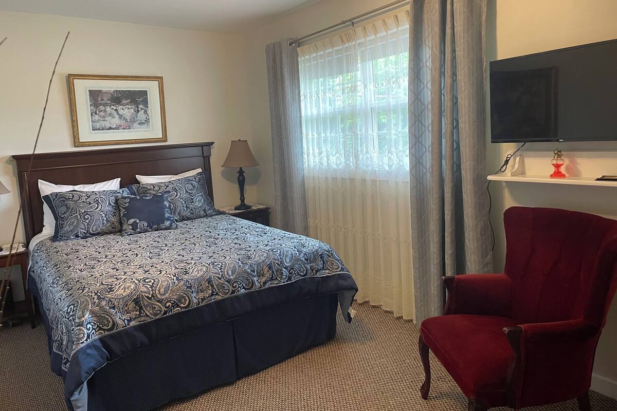A hotel room with a bed, a red armchair, a television, and a window with curtains.