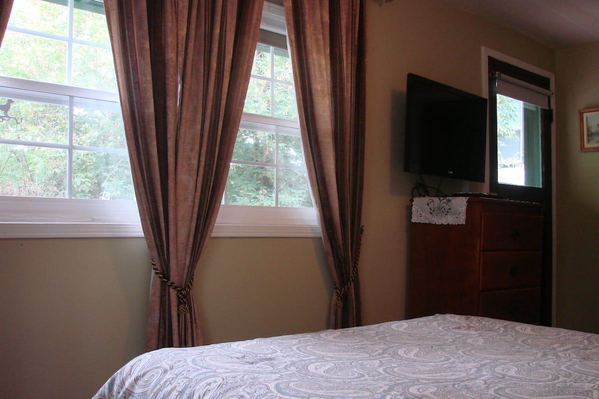 A bedroom with a bed, a dresser, a television on the wall, and a window with curtains.