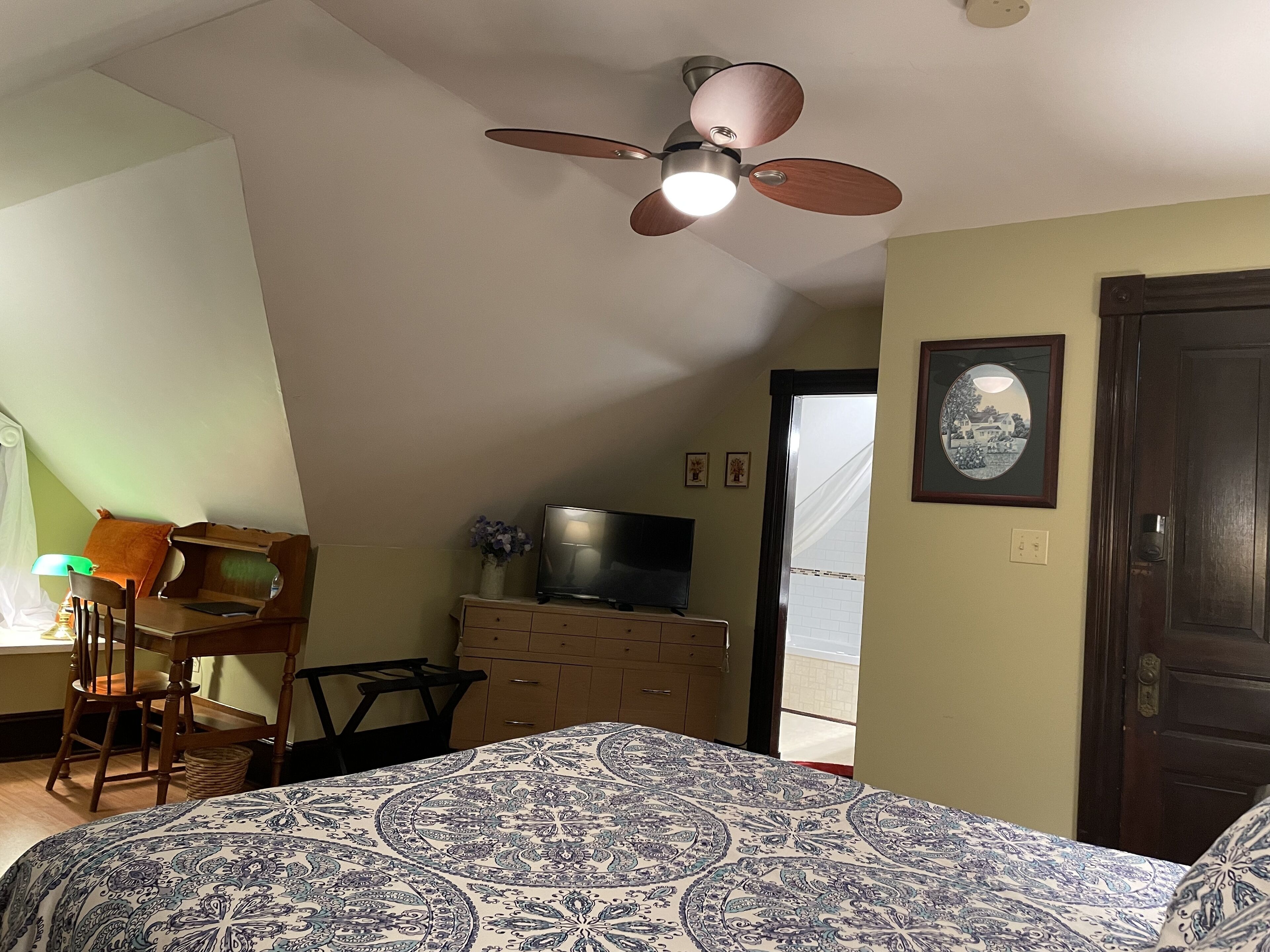 A bedroom with a bed, a desk, a chair, a dresser, a ceiling fan, and a door.