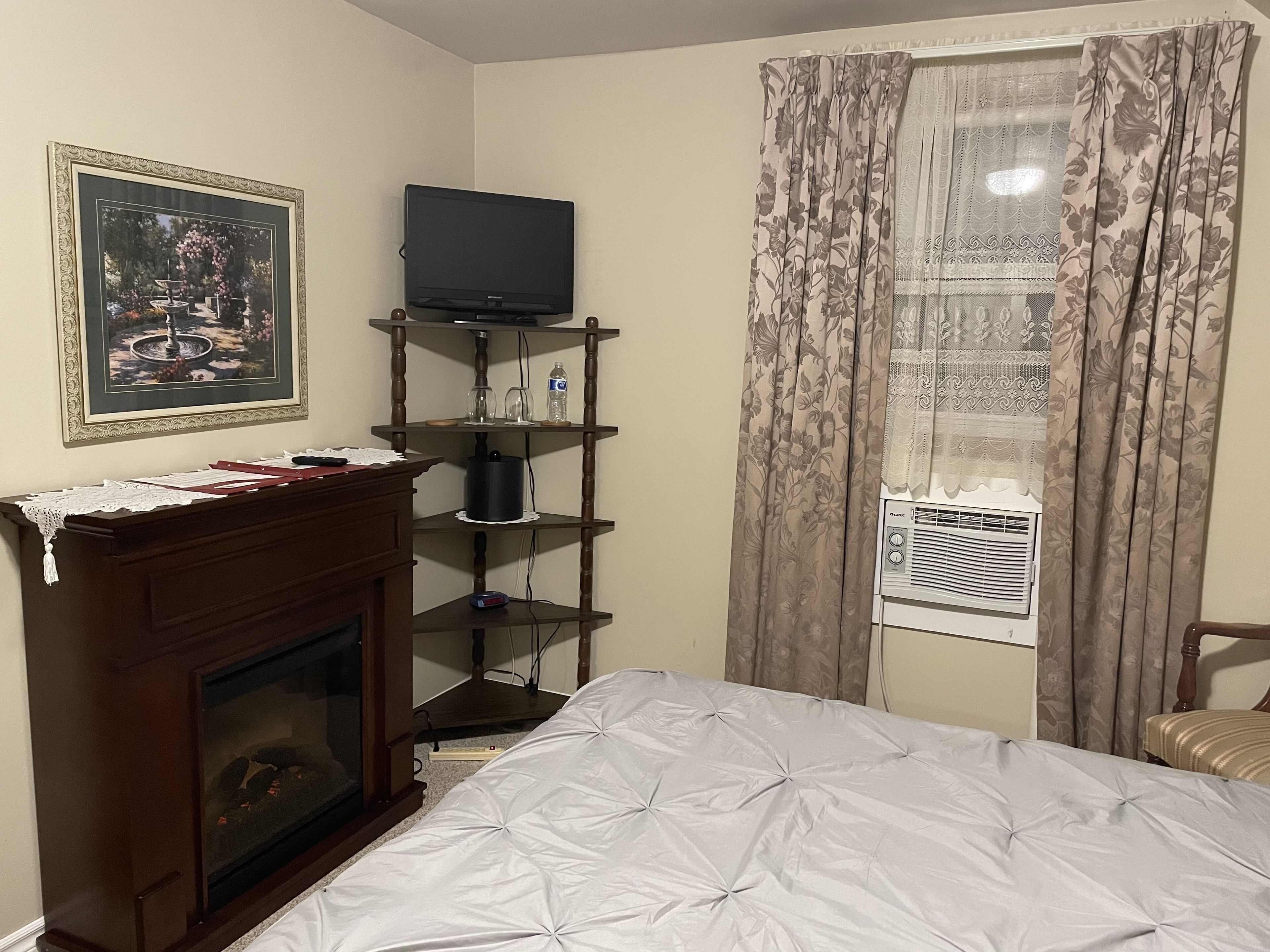 Une chambre à coucher avec un lit, un téléviseur posé sur une étagère en bois, une cheminée et une fenêtre avec des rideaux.