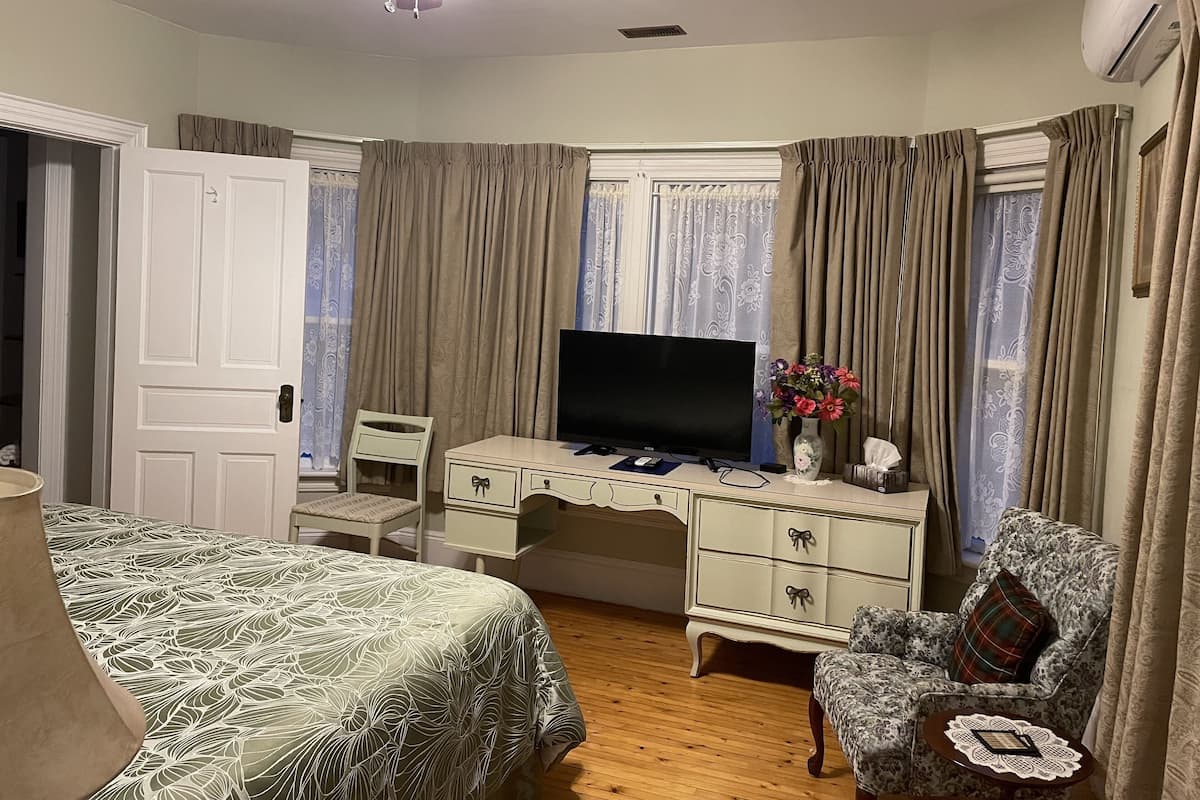 A bedroom with a bed, a desk with a TV, a chair, a dresser, and a window with curtains.