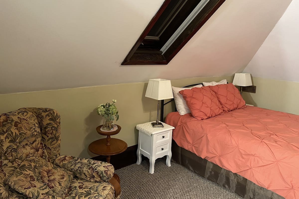 A small, cozy room with a bed, a chair, a nightstand, and a small table with a lamp.