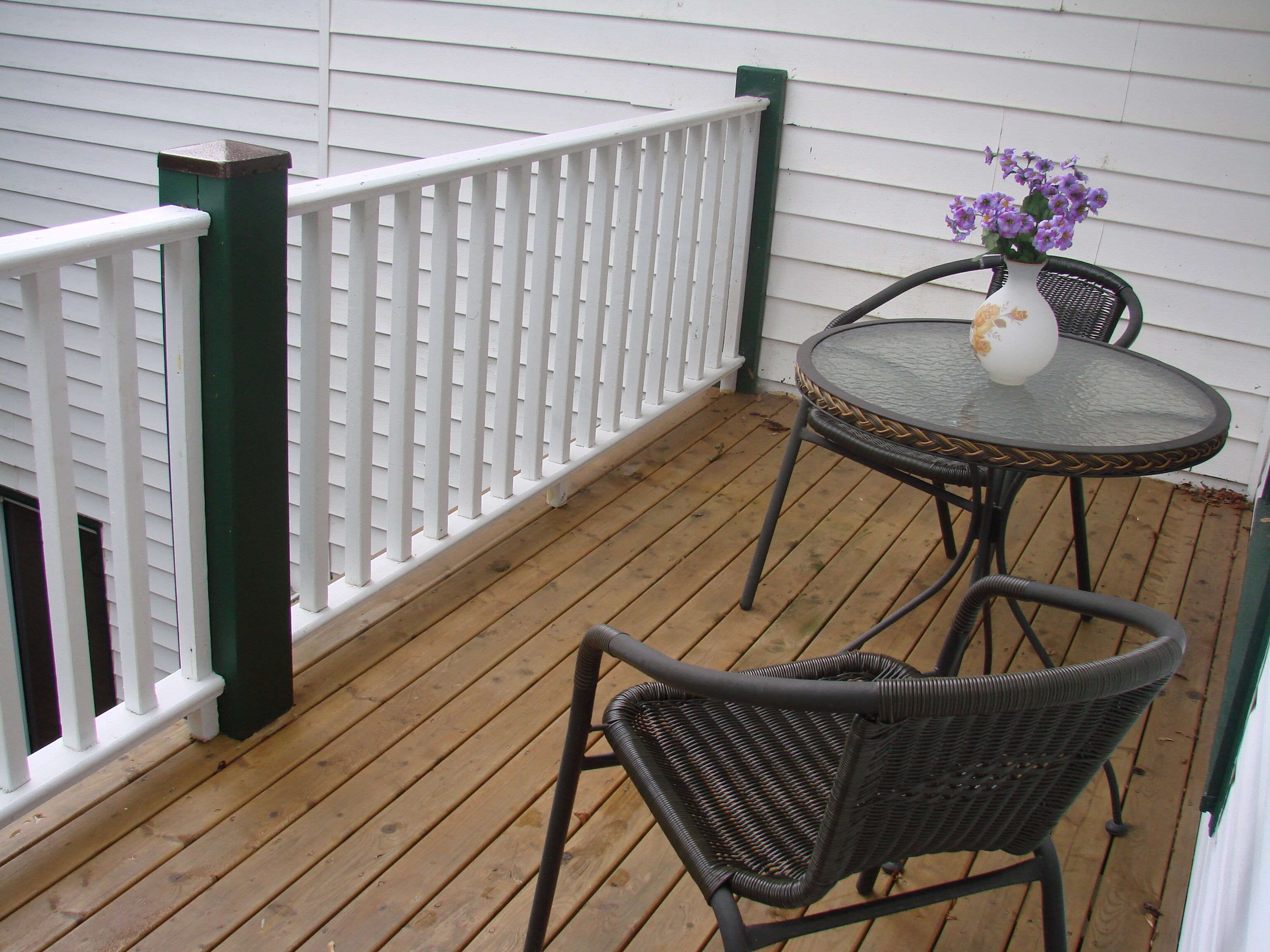 A wooden deck with a white railing and green posts, a round table with chairs, and a vase with purple flowers.