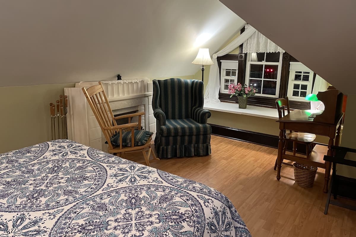 A bedroom with a large bed, a window with a view of the outside, a chair, a table, and a lamp.