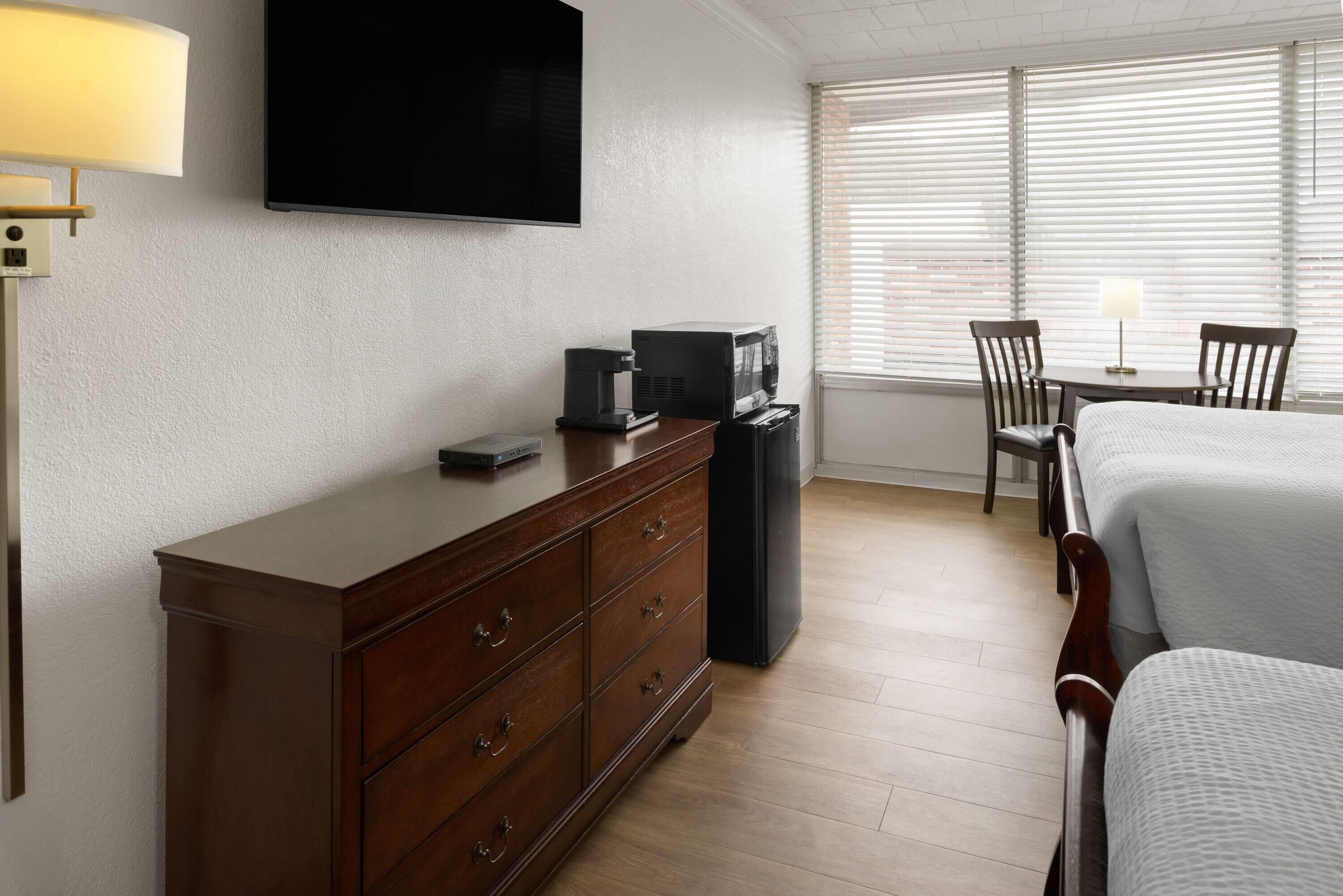 A hotel room with a bed, a desk with a coffee maker, a TV, and a window with blinds.