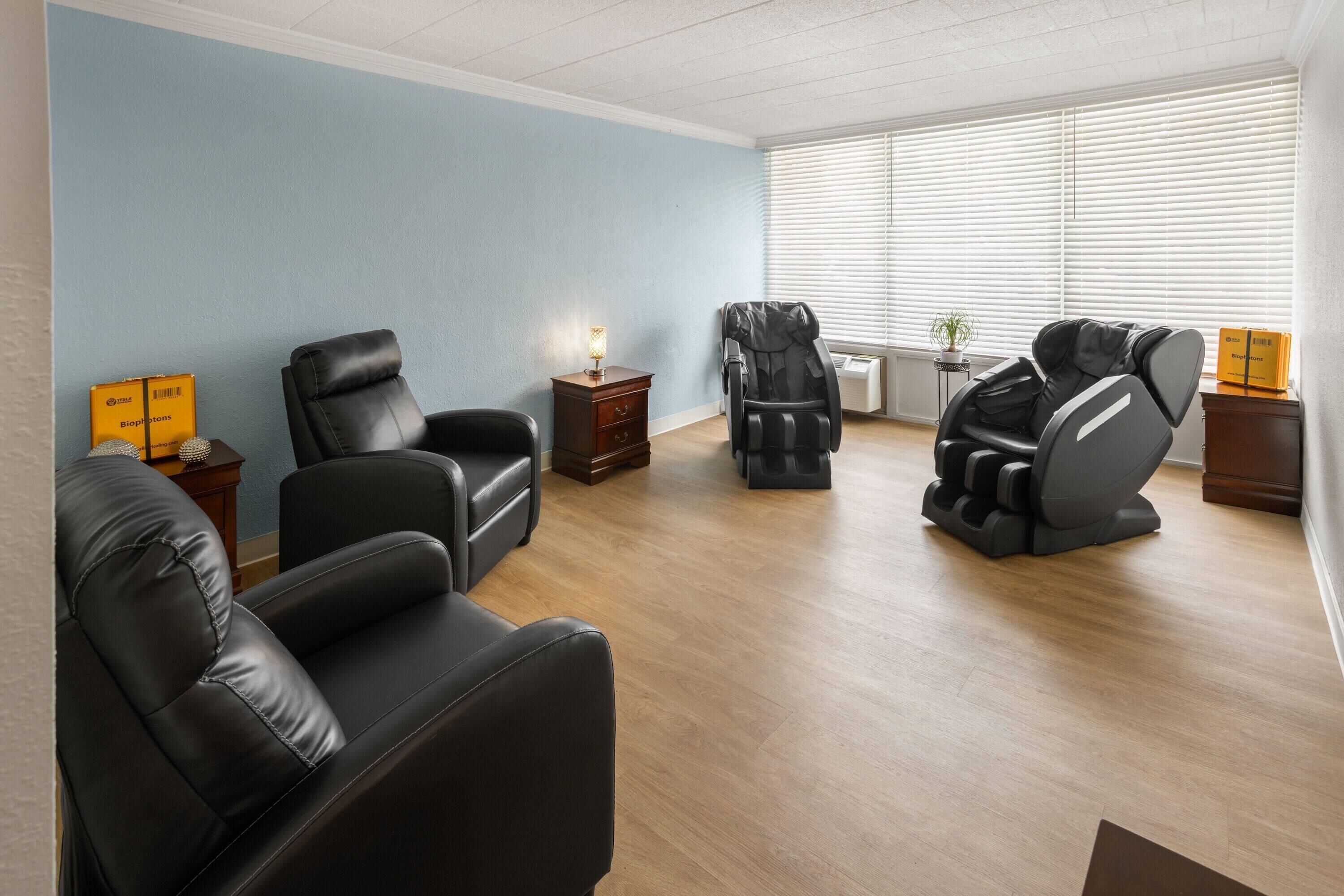 A room with a massage chair, a massage table, a small table with a lamp, and a window with blinds.