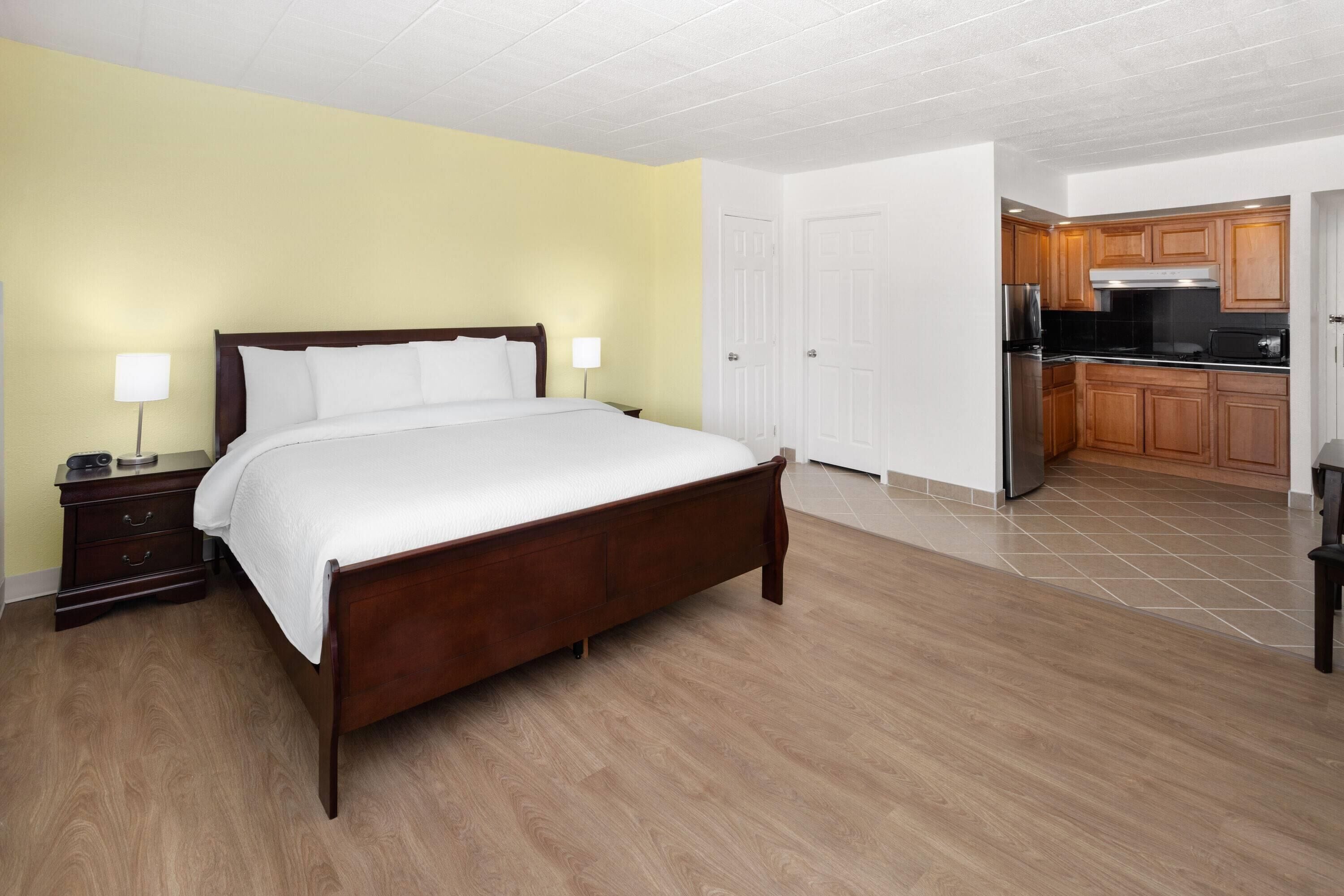 A bedroom with a wooden bed, a nightstand, a lamp, and a view into the kitchen.