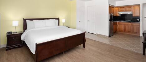 A bedroom with a wooden bed, a nightstand, a lamp, and a view into the kitchen.