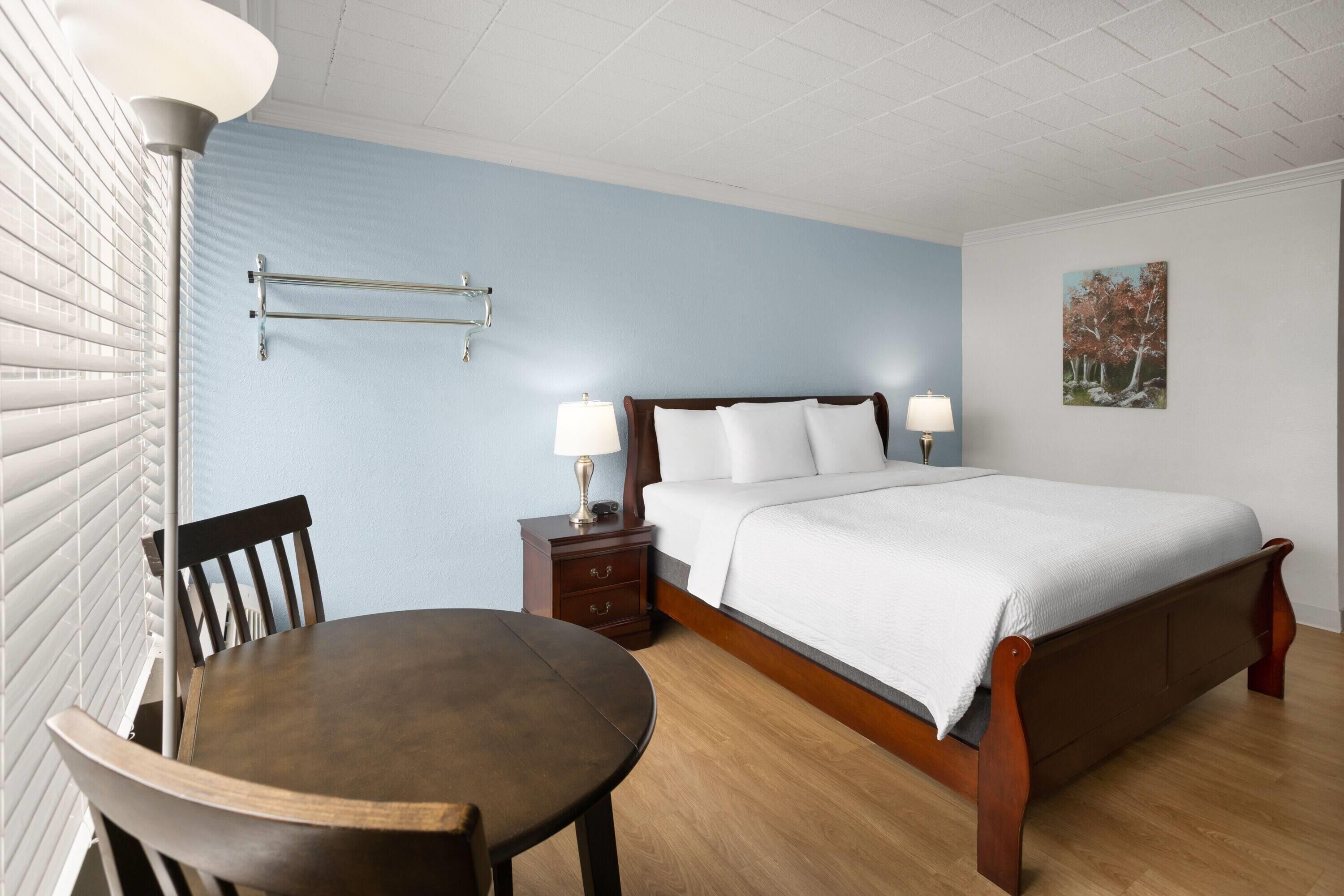 A bedroom with a bed, bedside table, lamp, chair, and a painting on the wall.