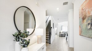 Un couloir avec une console blanche, un miroir rond, un lampadaire et un escalier menant à une autre pièce.
