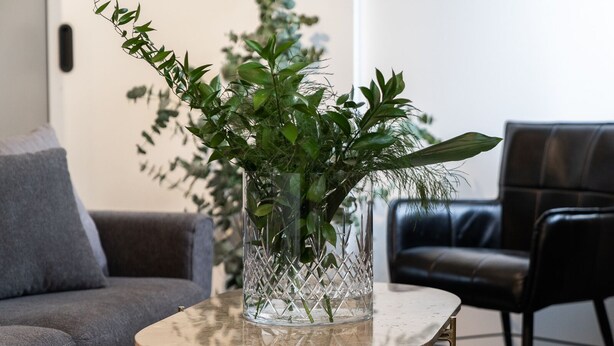 A living room with a grey sofa, a black leather chair, and a glass vase with green plants on a marble table. - Keflavik Luxury Apartments (Reykjanesbær)