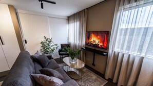 A modern living room with a grey sofa, a small side table with a plant, a flat-screen TV mounted on the wall, and a fireplace. - Keflavik Luxury Apartments (Reykjanesbær)