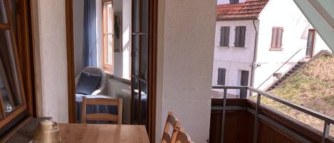 A wooden dining table with chairs, a view of a building through an open door, and a balcony with a railing.