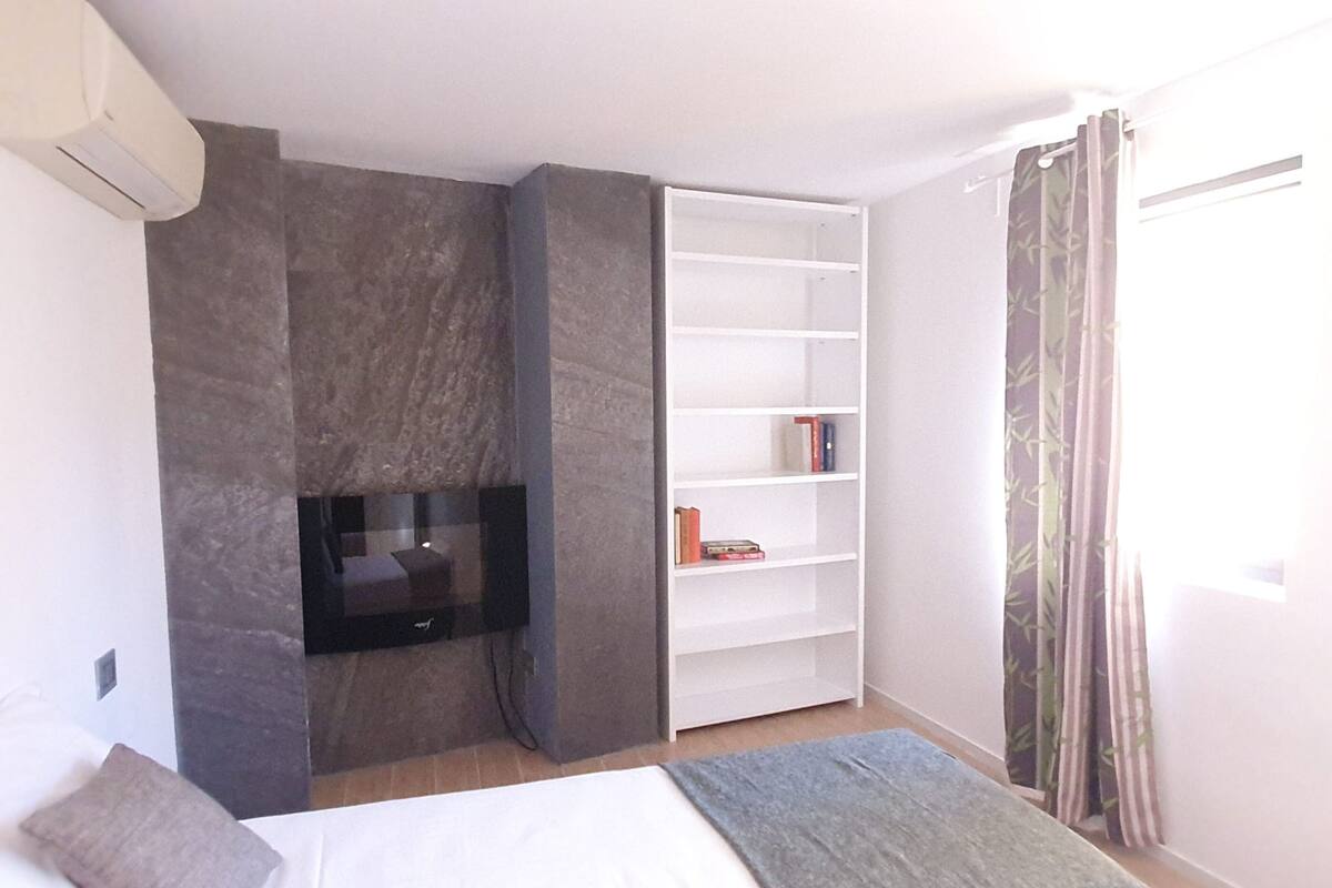 A bedroom with a bed, a TV, a bookshelf, and a window with curtains.