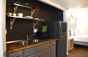 A compact kitchen with a refrigerator, microwave, and sink. There are wooden shelves above the counter holding dishes and a red cloth. A bed is visible in the adjacent room. - Montrose Pied-à-terre walking distance to Museums (Houston)