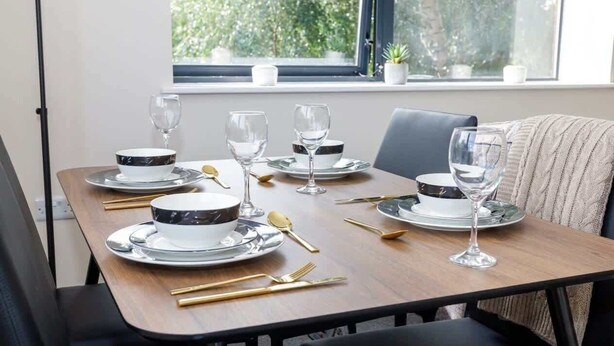 Una mesa de comedor dispuesta con platos blancos y negros, cubiertos dorados y copas de vino, ubicada cerca de una ventana con vistas a follaje.
