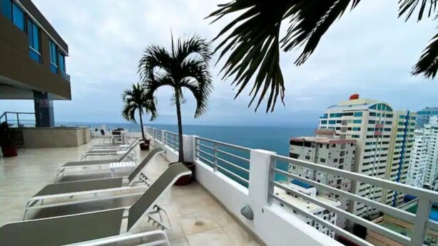 A balcony with metal chairs, a palm tree, and a view of the ocean and buildings.