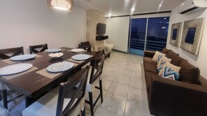A dining area with a table set for four, a sofa, a TV, and a balcony with a view.