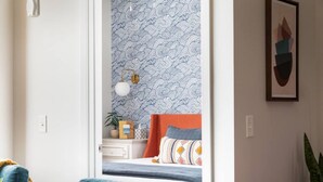 A bedroom with a patterned wallpaper, a bed with multiple pillows, a bedside table with a lamp, and a framed picture on the wall.