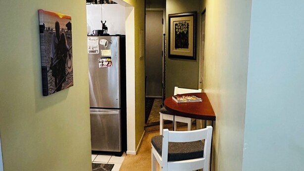 Un pasillo estrecho con un refrigerador, una mesa de comedor con sillas y un cuadro en la pared.