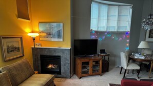Ein Wohnzimmer mit Kamin, Fernseher, einer Couch, einem roten Sessel und einem Esstisch mit Stühlen.