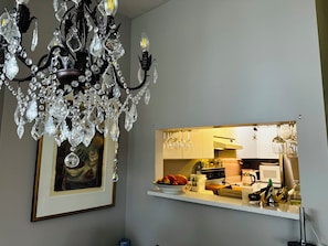 Um candelabro com detalhes em cristal, uma obra de arte emoldurada na parede e uma área de cozinha com bancada e diversos objetos.