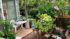 Ein überdachter Patio-Bereich mit Tisch und Stühlen, umgeben von Kübelpflanzen und einem kleinen Vogelhäuschen.
