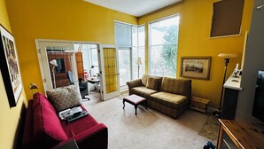 Ein Wohnzimmer mit einem roten Sofa, einem braunen Sofa und einem kleinen Tisch. Eine Tür führt in einen anderen Raum, und ein Fenster mit Jalousien ist vorhanden.