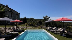 A rectangular swimming pool with clear blue water, surrounded by lounge chairs and umbrellas, with a stone building in the background.