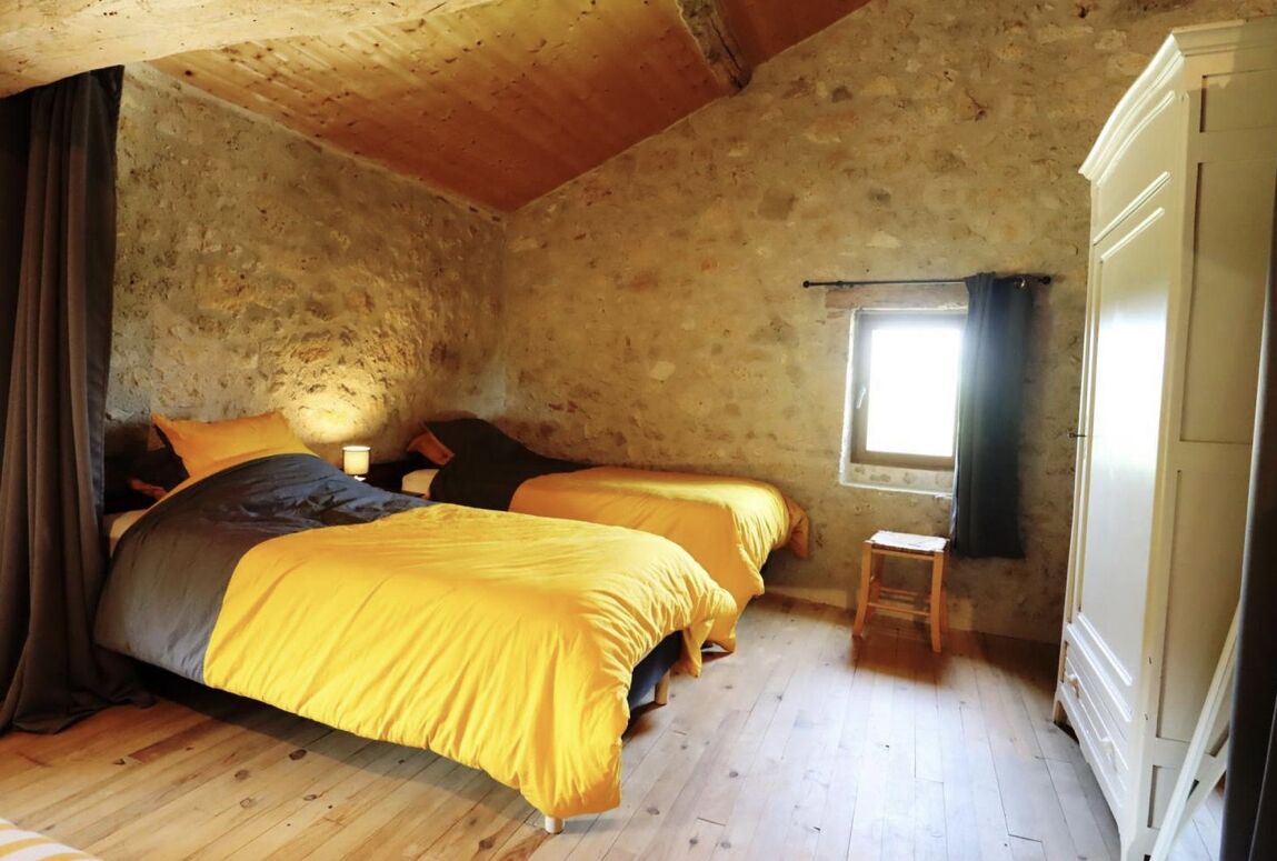 A rustic room with two beds, wooden floors, a small table, and a window with curtains.