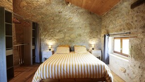 A bedroom with a stone wall, a wooden ceiling, a bed with striped bedding, bedside lamps, and a window with curtains.