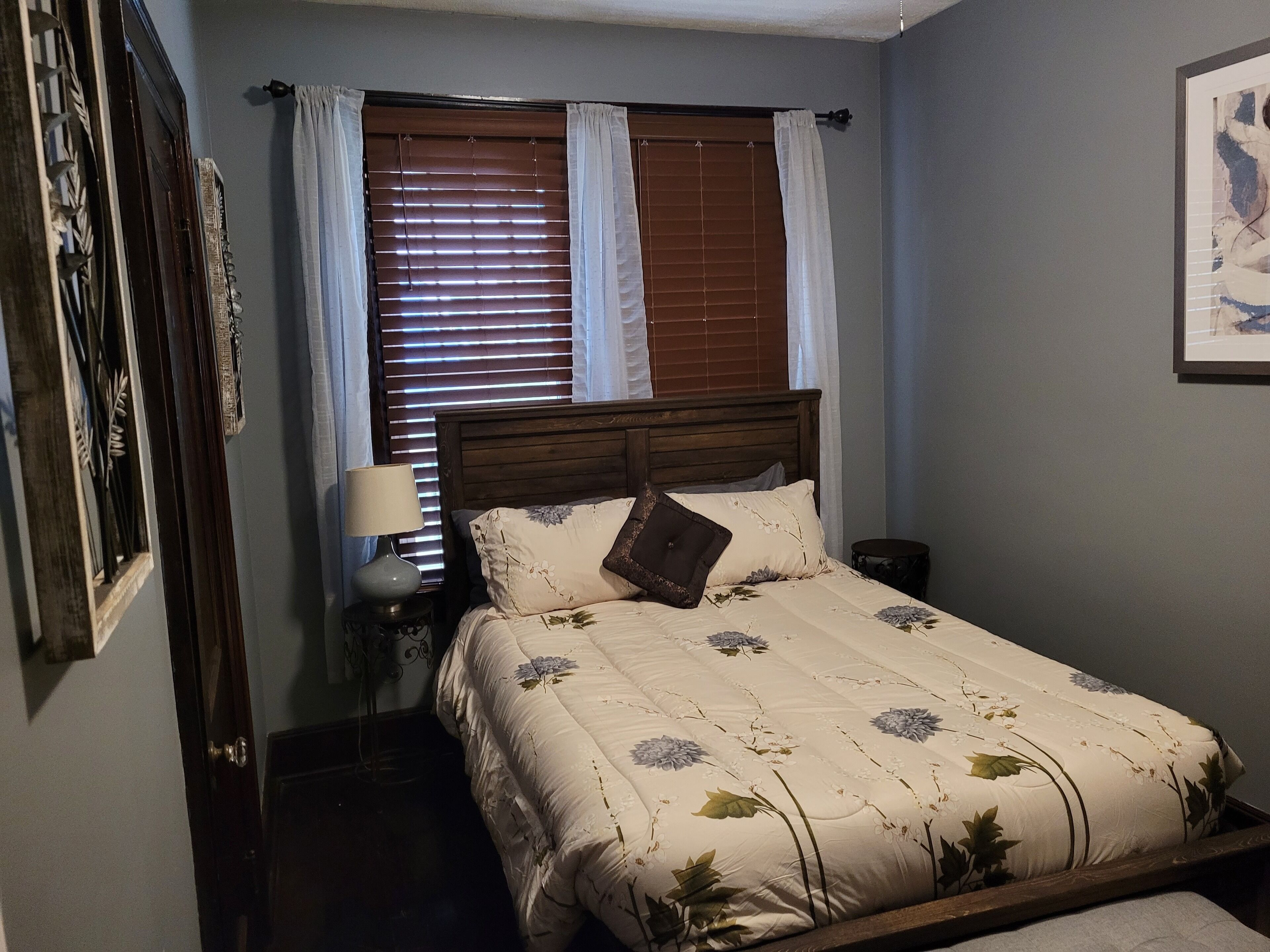 A bedroom with a wooden bed, a nightstand with a lamp, a window with blinds, and a framed picture on the wall.