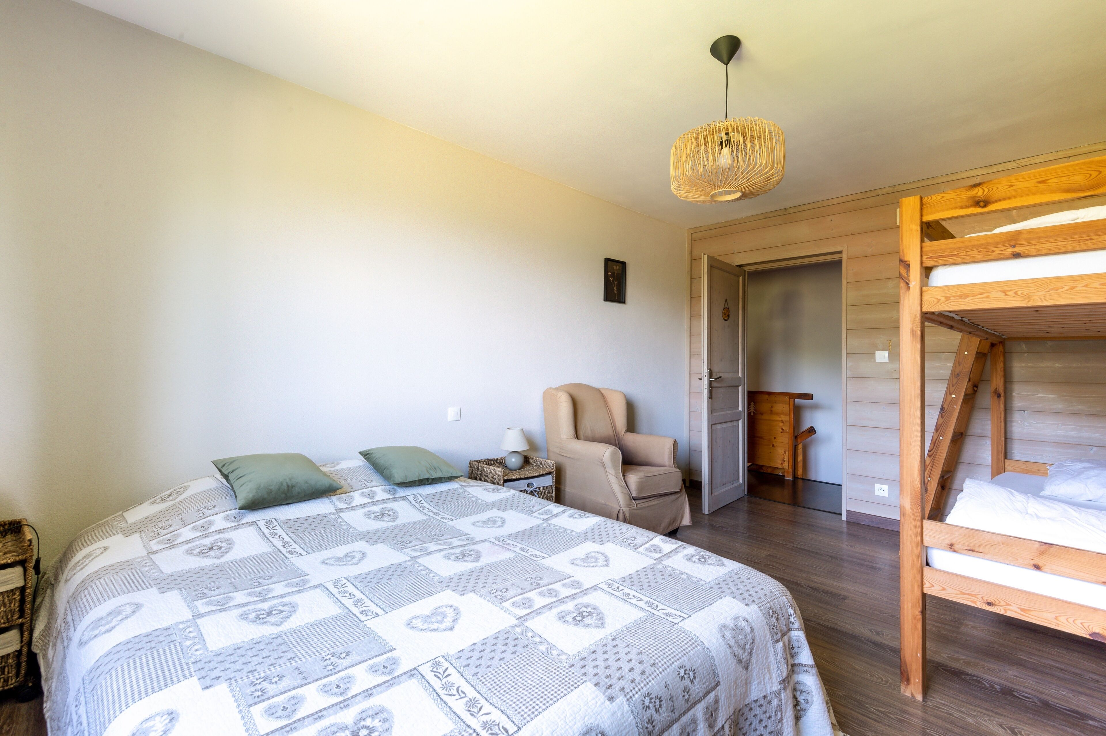 Ein Schlafzimmer mit einem großen Bett, einem Sessel, einem Schlafplatz in Etagenbauweise aus Holz, einer gewobenen Leuchte und einer Tür zu einem anderen Raum.