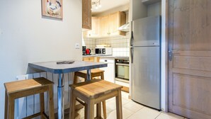 Een compacte keuken met houten kastjes, een roestvrijstalen koelkast, een magnetron en een koffiezetapparaat. Er is een kleine eettafel met twee krukken.