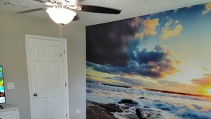 Une chambre à coucher avec un lit, un ventilateur de plafond, une porte blanche et un grand tableau représentant un coucher de soleil sur l’océan.