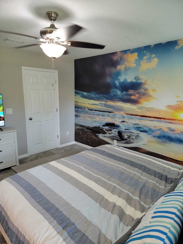 A bedroom with a bed, a ceiling fan, a white door, and a large painting of a sunset over the ocean.