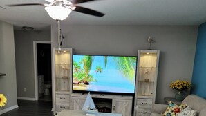 Un salon équipé d’un téléviseur à écran plat, d’un ventilateur de plafond, d’une table basse sur laquelle est posé un modèle de voilier, et d’un canapé.
