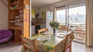 A dining area with a table set for two, wooden chairs, a wooden bookshelf, and a view of mountains through large windows. - Le Mirador - Studio avec balcon Vue Lac et Montagnes (Embrun)