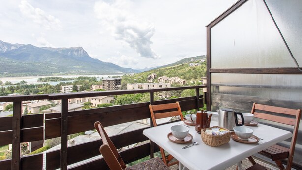 A balcony with a table set for two, overlooking a mountainous landscape and a river. - Le Mirador - Studio avec balcon Vue Lac et Montagnes (Embrun)