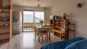 A room with a bed, a dining table with chairs, a refrigerator, a TV, and a bookshelf. - Le Mirador - Studio avec balcon Vue Lac et Montagnes (Embrun)