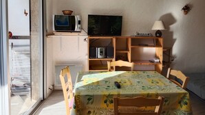 A dining area with a table covered in a Provence-themed tablecloth, a television on a shelf, a basket, and a lamp. - Le Mirador - Studio avec balcon Vue Lac et Montagnes (Embrun)