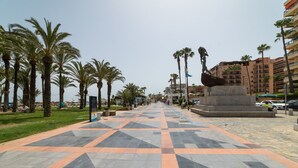 Un amplio paseo peatonal empedrado bordeado de palmeras y una estatua, que conduce a un conjunto de edificios.
