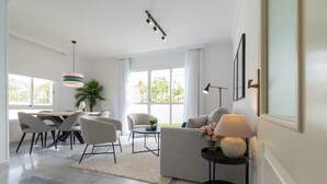 Una sala de estar moderna con un sofá, sillones, una mesa de comedor y una mesa de centro. Hay un ventanal con cortinas, una lámpara colgante y una planta pequeña sobre la mesa de centro.