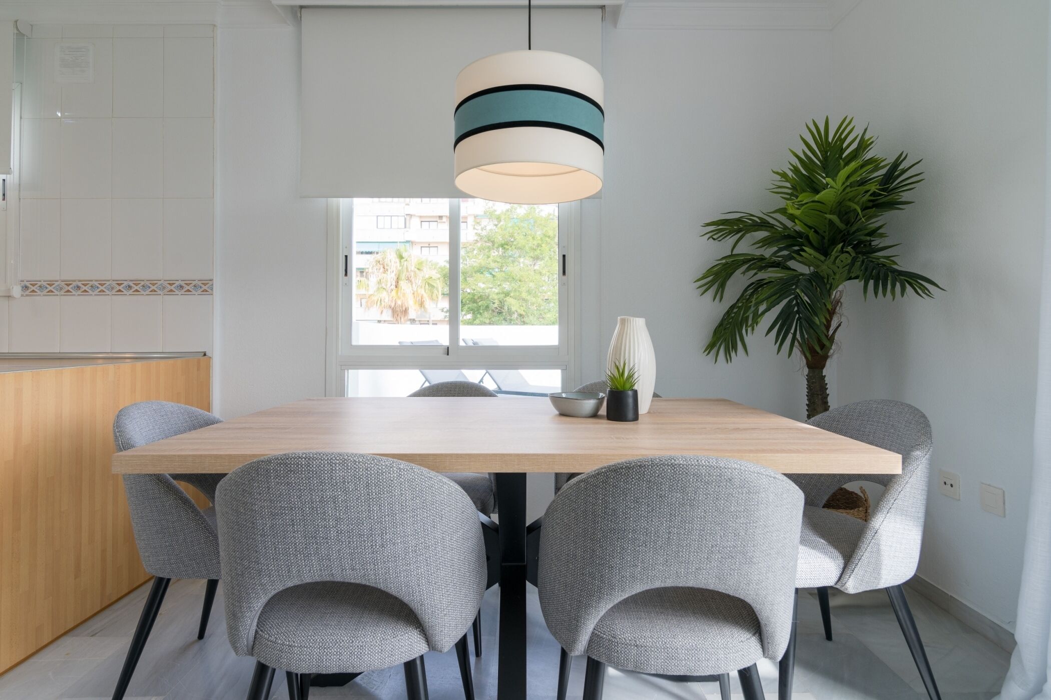 A modern dining area with a wooden table, grey upholstered chairs, a hanging light fixture, a potted plant, and a view through a window.