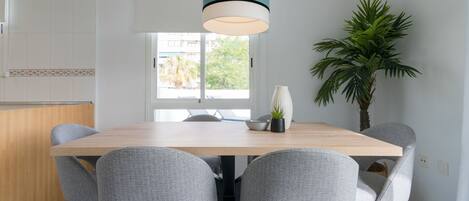 A modern dining area with a wooden table, grey upholstered chairs, a hanging light fixture, a potted plant, and a view through a window.