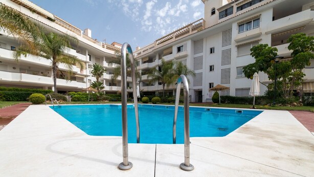 Una piscina con dos escaleras metálicas, rodeada por un edificio blanco con balcones y palmeras.