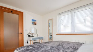 Un dormitorio con cama, una mesita de noche con espejo, cómoda y ventana con persianas.