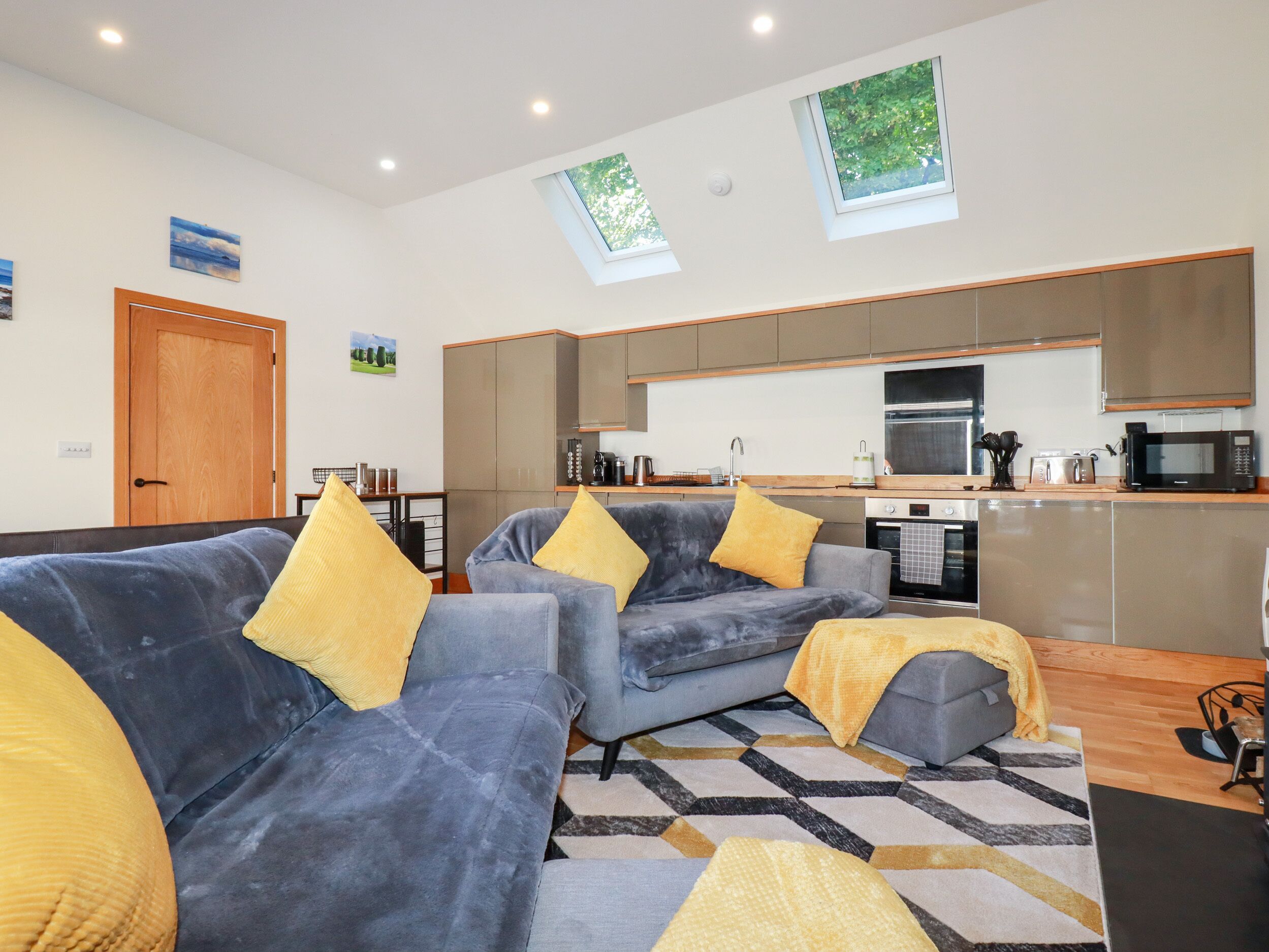 A modern living room with two grey sofas and yellow cushions, a kitchen with a microwave and oven, and a wooden door.