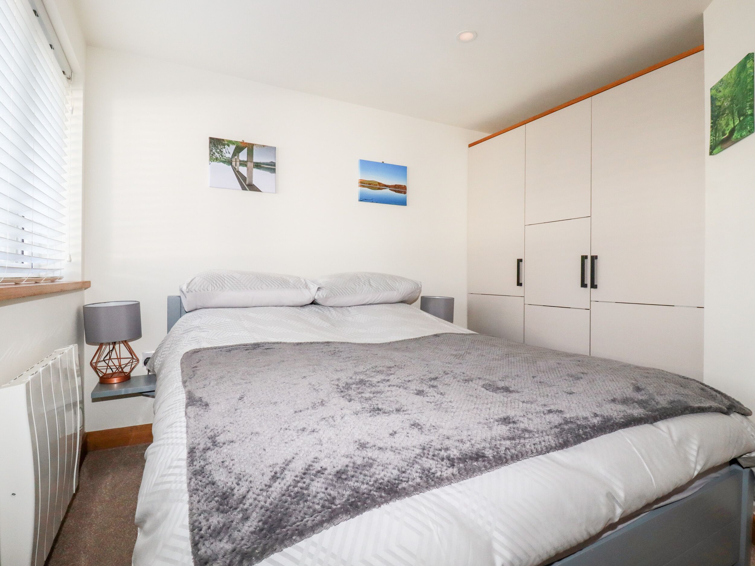 A bedroom with a bed, two wall-mounted pictures, a radiator, a lamp, and a wardrobe.