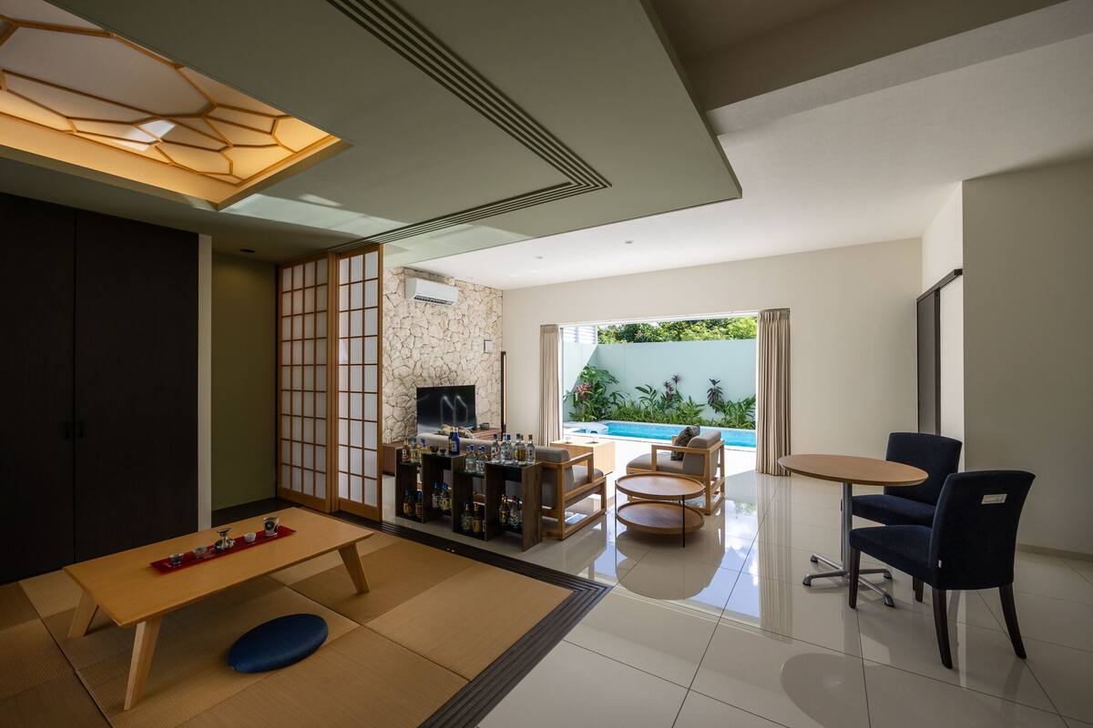 Una sala de estar moderna con una mesa de madera, sofá y televisor, una pared de piedra y vista a una piscina.